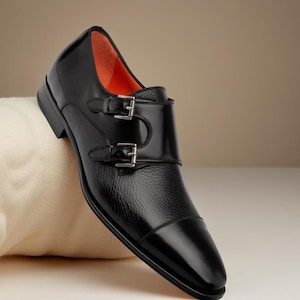 May include: A black leather double monk strap shoe with a textured vamp and a smooth, glossy toe cap. The shoe has two silver buckles and a bright orange interior lining. The shoe is set against a neutral background.