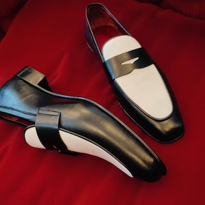 May include: A pair of black and white leather loafers. The shoes feature a classic design with a black upper, a white vamp, and a black strap. The shoes are set against a red background.