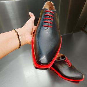 May include: Black leather dress shoes with red soles and laces. The shoes have a sleek, modern design with a pointed toe. The red accents provide a striking contrast to the black leather. The shoes are displayed on a reflective surface.