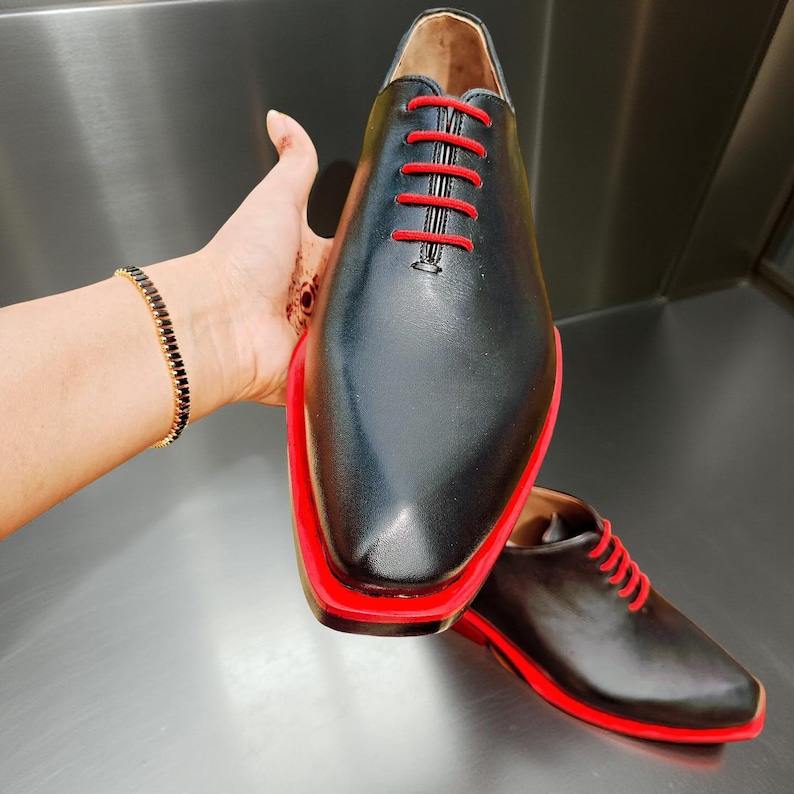 May include: Black leather dress shoes with red soles and laces. The shoes have a sleek, modern design with a pointed toe. The red accents provide a striking contrast to the black leather. The shoes are displayed on a reflective surface.