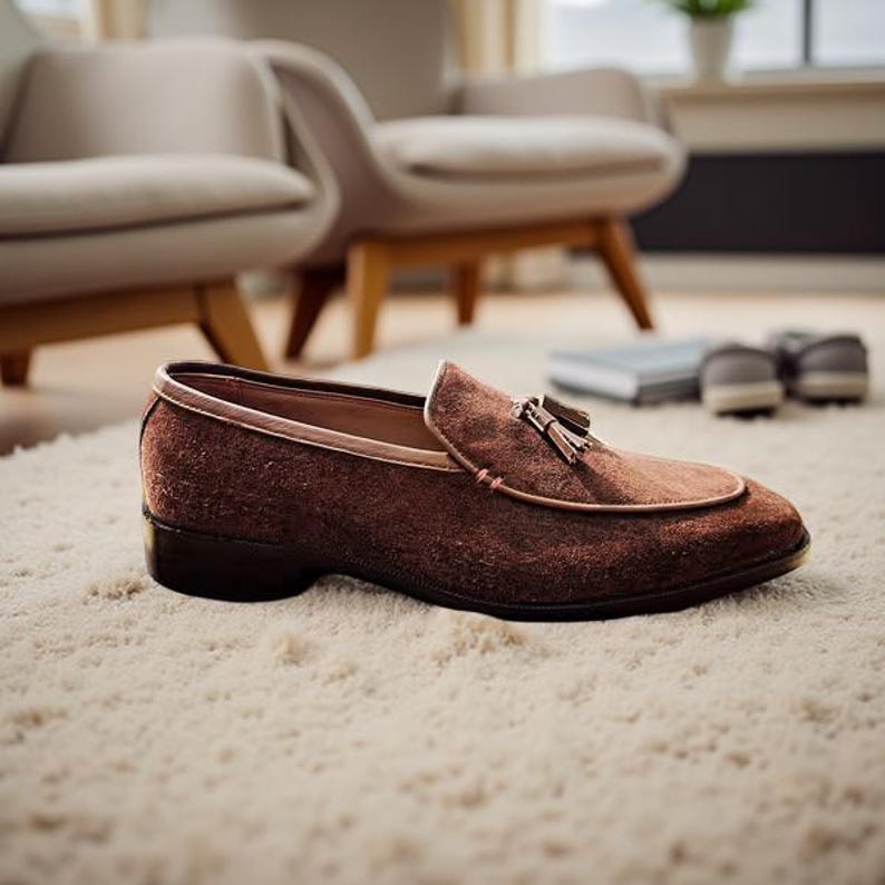 May include: A brown suede loafer with a tassel detail, set on a cream-colored rug. The shoe has a leather trim and a dark sole. The background includes two chairs and a window.