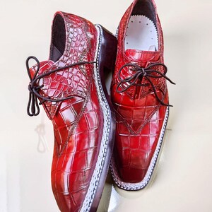 May include: A pair of red leather dress shoes with a textured, crocodile-like pattern. The shoes have dark brown laces, a white trim around the sole, and a dark brown heel. The shoes are displayed on a light-colored surface.