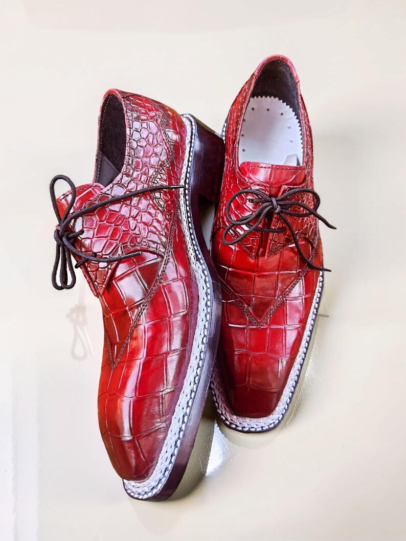 May include: A pair of red leather dress shoes with a textured, crocodile-like pattern. The shoes have dark brown laces, a white trim around the sole, and a dark brown heel. The shoes are displayed on a light-colored surface.