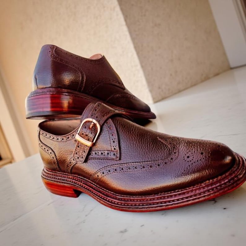 K&ouml;nnte beinhalten: Ein Paar braune Lederschuhe mit Schnallenverschluss und dekorativen Perforationen. Die Schuhe haben eine polierte Oberfl&auml;che und eine rot get&ouml;nte Sohle. Ein Schuh ist auf dem anderen positioniert.
