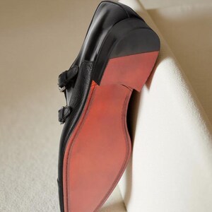 May include: Black leather double monk strap shoe with a red sole. The shoe has two silver buckles and a pointed toe. The sole is a vibrant red, contrasting with the black leather upper. The shoe is leaning against a white surface.