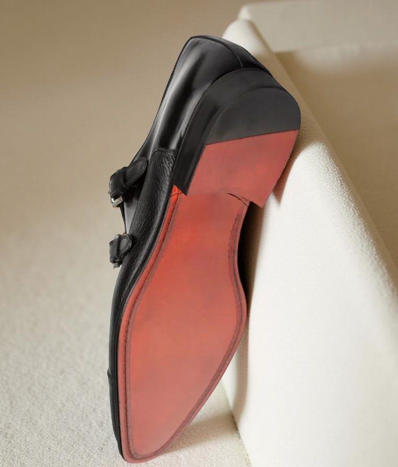 May include: Black leather double monk strap shoe with a red sole. The shoe has two silver buckles and a pointed toe. The sole is a vibrant red, contrasting with the black leather upper. The shoe is leaning against a white surface.