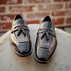 May include: A pair of black leather loafers with a crocodile texture. The shoes have a tassel detail and a rounded toe. The soles are dark with a white trim. The shoes are displayed on a light-colored surface.