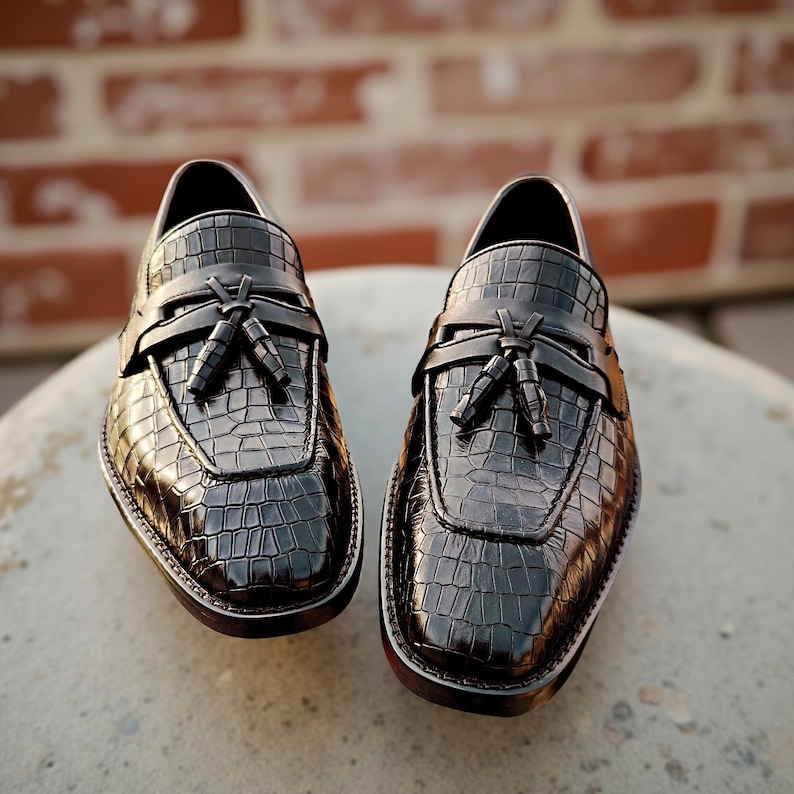 May include: A pair of black leather loafers with a crocodile texture. The shoes have a tassel detail and a rounded toe. The soles are dark with a white trim. The shoes are displayed on a light-colored surface.