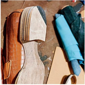May include: Close-up of a partially constructed brown leather shoe with a crocodile pattern. The sole is unfinished, revealing layers of materials. A light blue fabric and other materials are in the background, suggesting a workshop setting.