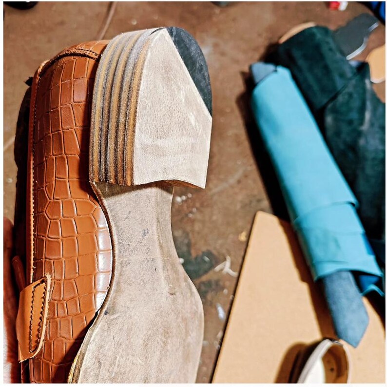 May include: Close-up of a partially constructed brown leather shoe with a crocodile pattern. The sole is unfinished, revealing layers of materials. A light blue fabric and other materials are in the background, suggesting a workshop setting.