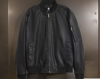 May include: A black faux leather bomber jacket with a zipper closure and a small pocket on the left sleeve. The jacket has a ribbed collar and cuffs.