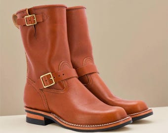 May include: A pair of brown leather boots with a square toe and a mid-calf height. The boots feature gold-colored buckles and straps, with a stitched sole. The boots are approximately 10 inches tall.