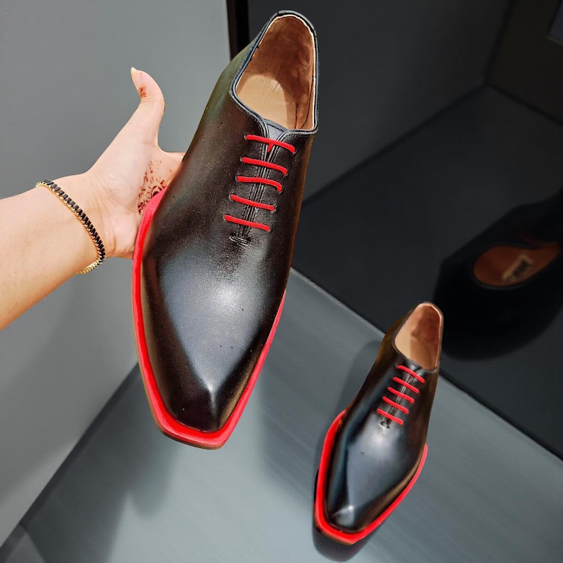 May include: Black leather Oxford shoes with red soles and laces. The shoes have a classic design with a pointed toe. The image shows a pair of shoes, one held up and the other on a reflective surface.