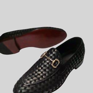 May include: Black woven leather loafers with a gold-colored metal bit detail. The shoes have a classic slip-on design and a red sole. The image shows one shoe in focus and another in the background.
