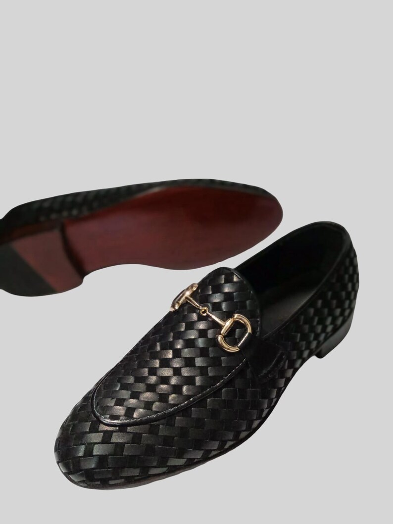 May include: Black woven leather loafers with a gold-colored metal bit detail. The shoes have a classic slip-on design and a red sole. The image shows one shoe in focus and another in the background.