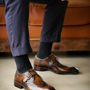 May include: Brown leather dress shoes with a buckle closure and brogue detailing. The shoes are worn with black socks and navy pinstripe trousers. The shoes have a polished finish and a dark brown sole.