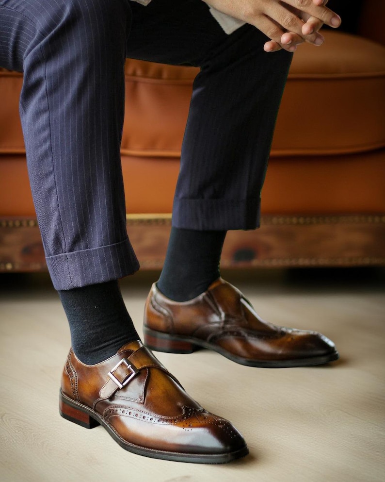 May include: Brown leather dress shoes with a buckle closure and brogue detailing. The shoes are worn with black socks and navy pinstripe trousers. The shoes have a polished finish and a dark brown sole.