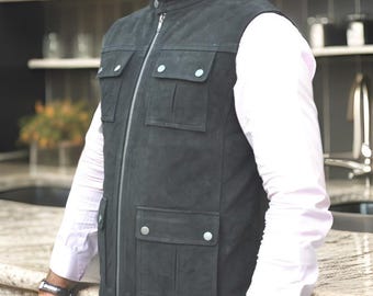 May include: A charcoal gray suede vest with a stand-up collar and a silver zipper. The vest features four front pockets with button closures. The model is wearing a white dress shirt underneath the vest.