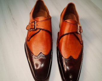 May include: A pair of two-tone leather shoes with a buckle fastening. The shoes are brown and dark brown with decorative perforations along the toe cap. The shoes have a square toe and a leather sole.