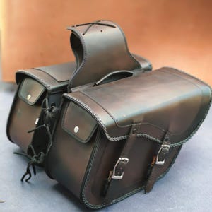 May include: A pair of black leather motorcycle saddlebags with buckles and stitching. The bags are attached to a leather strap with a buckle.
