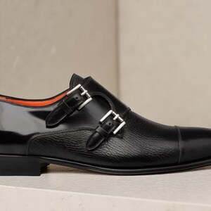 May include: Black leather double monk strap shoe with a glossy finish on the heel and toe. The shoe features two silver buckles and a textured leather midsection. The interior lining is orange.