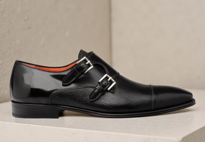 May include: Black leather double monk strap shoe with a glossy finish on the heel and toe. The shoe features two silver buckles and a textured leather midsection. The interior lining is orange.