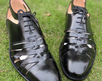 May include: A pair of black leather lace-up shoes featuring a woven cage design across the vamp and sides, a closed toe, and an open heel construction with a thin ankle strap. The shoes have a polished finish and black laces tied at the top.