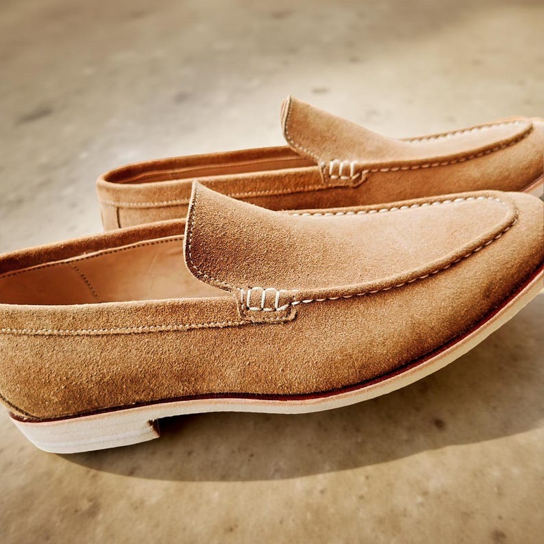 May include: A pair of tan suede loafers. The shoes feature a classic slip-on design with a rounded toe and a low heel. White stitching accents the seams and the top of the shoe. The sole is white with a thin red stripe.