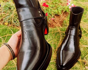 May include: Black leather boots with a square toe and a strap across the ankle. The boots have a stacked platform sole and a red accent on the heel. One boot is held up, and another boot is on the ground.