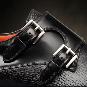 May include: Detail of a black leather shoe featuring two silver buckles. The shoe has a textured finish and a smooth leather strap. The buckles are rectangular and polished, contrasting with the matte leather.