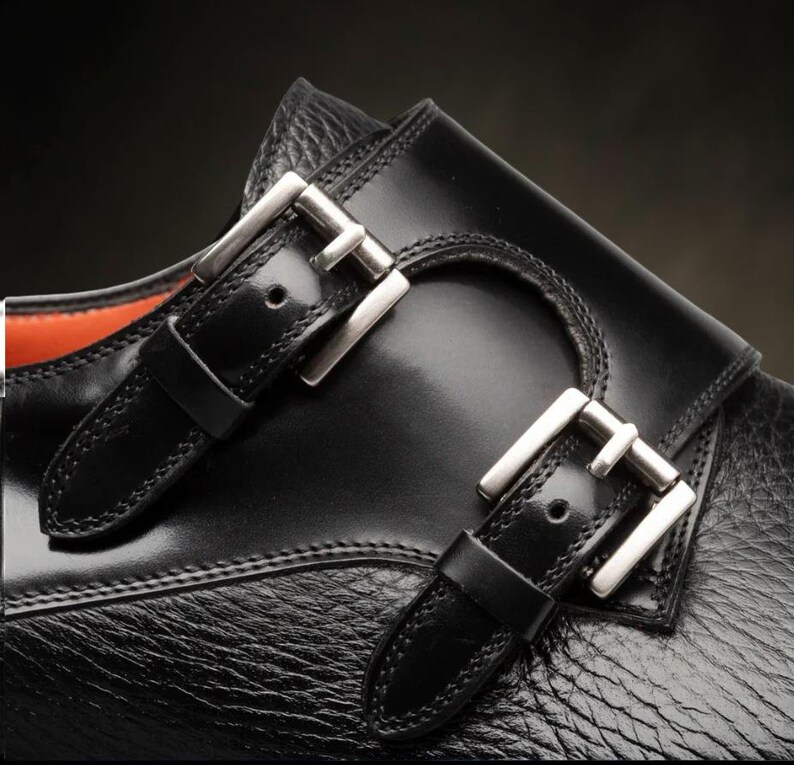 May include: Detail of a black leather shoe featuring two silver buckles. The shoe has a textured finish and a smooth leather strap. The buckles are rectangular and polished, contrasting with the matte leather.
