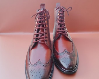 May include: A pair of burgundy leather boots with brogue detailing. The boots have a lace-up design with matching burgundy laces and eyelets. The leather has a polished finish, and the boots are set against a solid background.