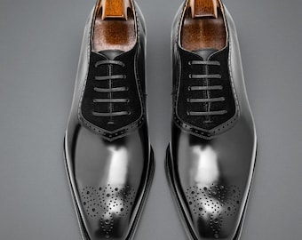 May include: A pair of black leather dress shoes with a glossy finish. The shoes feature a suede panel on the vamp, decorative perforations, and wooden shoe trees. The laces are black, and the shoes have a square toe.