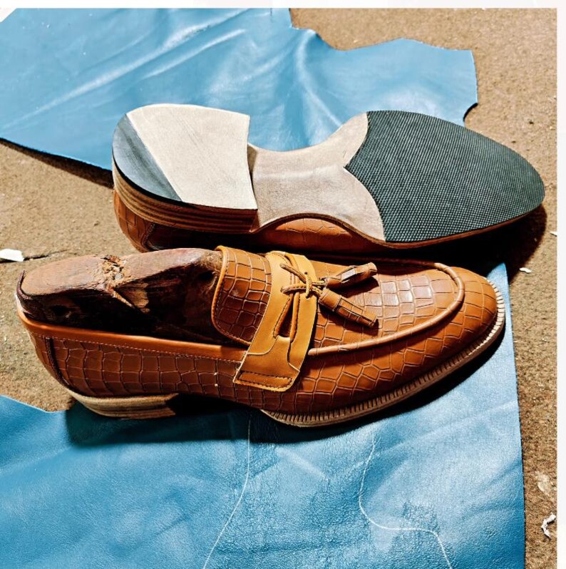 May include: Two brown leather shoes, one complete and one in the process of being made, sit on a blue surface. The finished shoe has a crocodile texture, a strap with a tassel, and a light brown sole. The unfinished shoe shows the layers of construction.