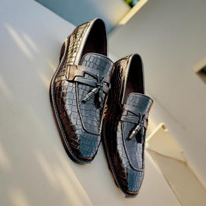 May include: Pair of glossy black leather loafers with a crocodile texture. Each shoe features a decorative strap with a tassel. The shoes are angled, showcasing their design and craftsmanship.