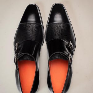 May include: A pair of black leather dress shoes with a textured midsection and silver buckles. The shoes have a glossy finish and orange insoles, showcasing a classic design.