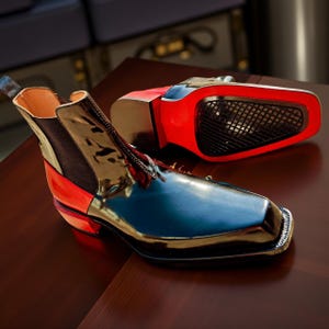 May include: A pair of high-gloss, square-toe Chelsea boots. The boots feature a black and blue upper with red accents on the heel and sole. The sole is red with a black mesh detail. The boots are displayed on a dark brown surface.