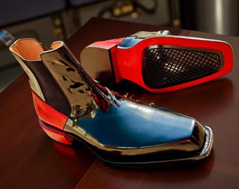 May include: A pair of high-gloss, square-toe Chelsea boots. The boots feature a black and blue upper with red accents on the heel and sole. The sole is red with a black mesh detail. The boots are displayed on a dark brown surface.