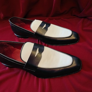 May include: A pair of black and white leather loafers. The shoes feature a classic design with a white vamp and a black strap. The interior is red. The shoes are displayed on a red velvet surface.