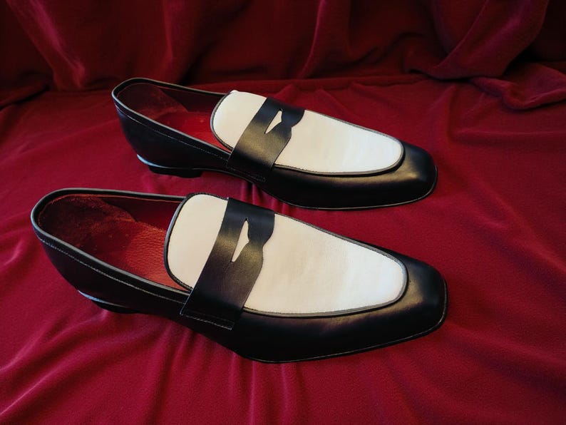 May include: A pair of black and white leather loafers. The shoes feature a classic design with a white vamp and a black strap. The interior is red. The shoes are displayed on a red velvet surface.