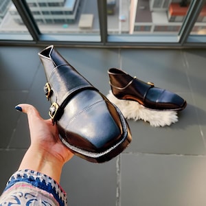 Pu&ograve; includere: Scarpe in pelle nera con punta quadrata e fibbie color oro. Le scarpe hanno un design unico con un cinturino sulla parte superiore e una caviglia bassa. Una scarpa &egrave; tenuta in mano, l'altra poggia su una superficie bianca e soffice.