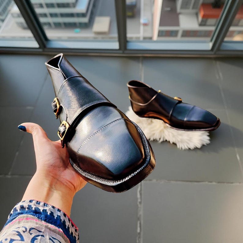 Pu&ograve; includere: Scarpe in pelle nera con punta quadrata e fibbie color oro. Le scarpe hanno un design unico con un cinturino sulla parte superiore e una caviglia bassa. Una scarpa &egrave; tenuta in mano, l'altra poggia su una superficie bianca e soffice.