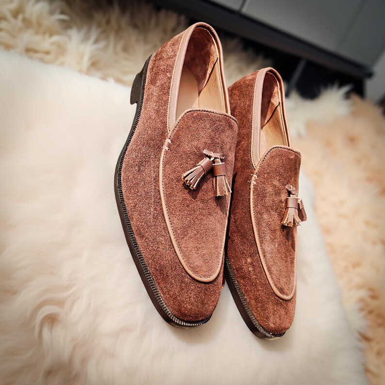 May include: A pair of brown suede loafers with tassel details. The shoes have a rounded toe and a stitched sole. The interior lining is a lighter shade of brown. The loafers are displayed on a white, fluffy surface.