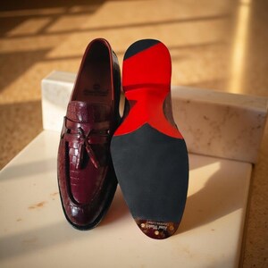 May include: Burgundy leather loafers with a crocodile texture, featuring a tassel detail and a black sole with a red accent. The shoes are displayed on a white surface, showcasing the craftsmanship and design.