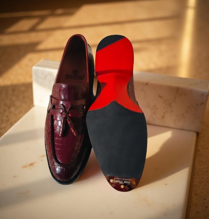 May include: Burgundy leather loafers with a crocodile texture, featuring a tassel detail and a black sole with a red accent. The shoes are displayed on a white surface, showcasing the craftsmanship and design.