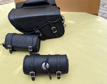 May include: A set of three black motorcycle bags including one large saddlebag with two buckle straps and two cylindrical tool bags. One tool bag features an embossed eagle design, while the other has a central silver-toned circular concho.