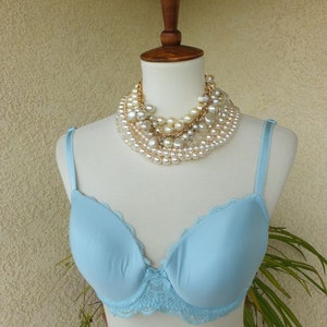 May include: A light blue bra with lace trim on a white mannequin. The bra has a smooth, seamless cup and adjustable straps. The mannequin is wearing a multi-strand pearl necklace.