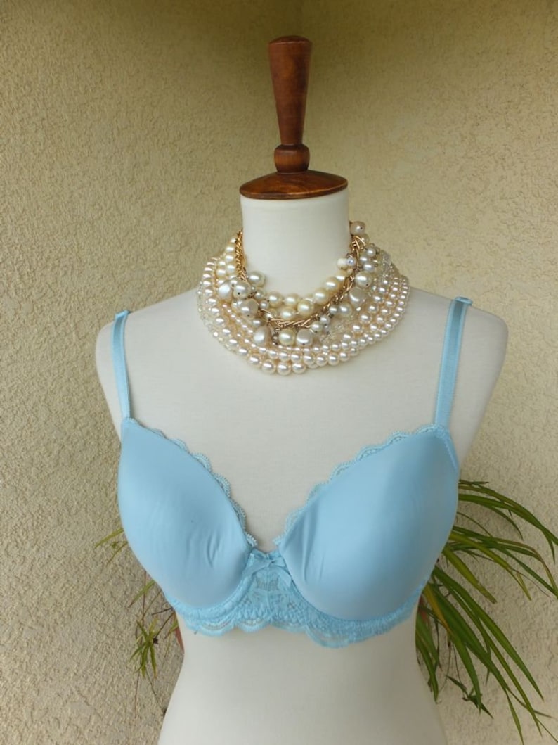May include: A light blue bra with lace trim on a white mannequin. The bra has a smooth, seamless cup and adjustable straps. The mannequin is wearing a multi-strand pearl necklace.