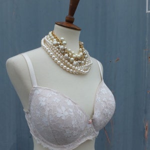 K&ouml;nnte beinhalten: Ein wei&szlig;er Spitzen-BH mit zarten Blumenmustern auf einer wei&szlig;en Schaufensterpuppe. Die Schaufensterpuppe tr&auml;gt mehrere Perlen- und Goldketten.
