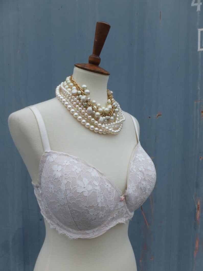 K&ouml;nnte beinhalten: Ein wei&szlig;er Spitzen-BH mit zarten Blumenmustern auf einer wei&szlig;en Schaufensterpuppe. Die Schaufensterpuppe tr&auml;gt mehrere Perlen- und Goldketten.